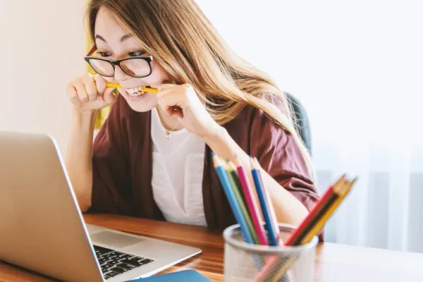 Une jeune femme assise en face d'un MacBook, mord dans un crayon jaune.